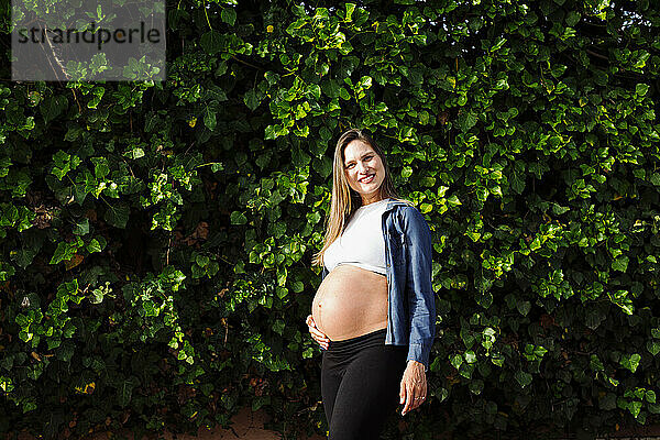 Lächelnde schwangere Frau steht vor einer Hecke