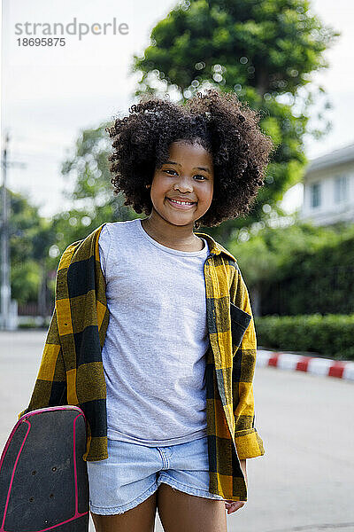 Smiling girl standing with skateboard on footpath