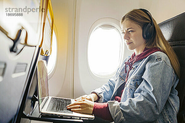 Junge Frau benutzt Laptop und hört Musik im Flugzeug