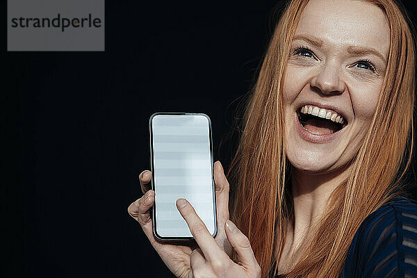 Junge Frau lacht und hält Smartphone vor schwarzem Hintergrund