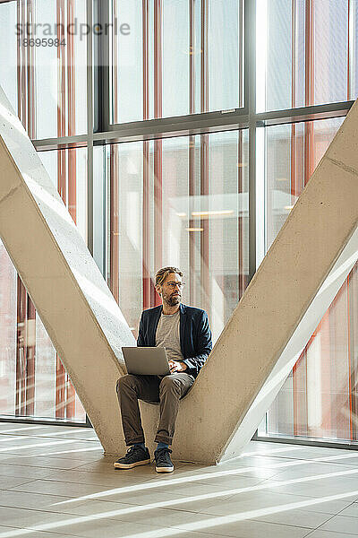 Geschäftsmann mit Laptop sitzt auf V-förmiger Säule im Gebäude