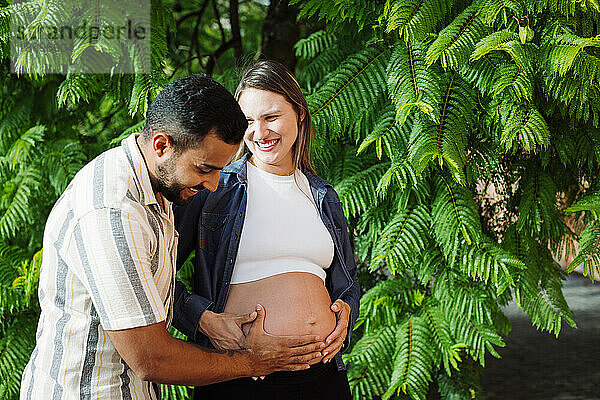 Smiling man touching belly of pregnant woman in front of tree