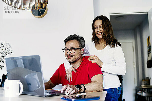 Frau mit Arm um Mann  der zu Hause am Laptop arbeitet