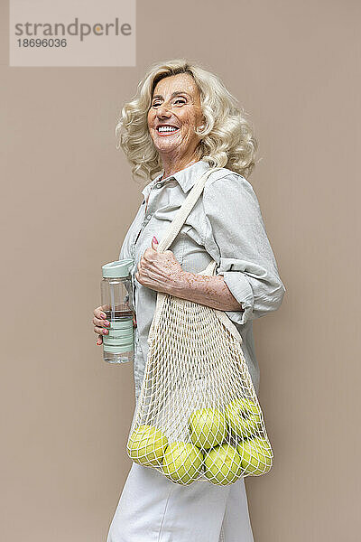 Cheerful senior businesswoman carrying apples in mesh bag against beige background