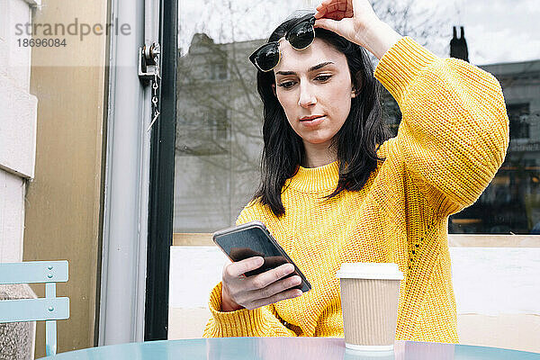 Frau im gelben Pullover hebt Sonnenbrille hoch und benutzt Smartphone im Straßencafé