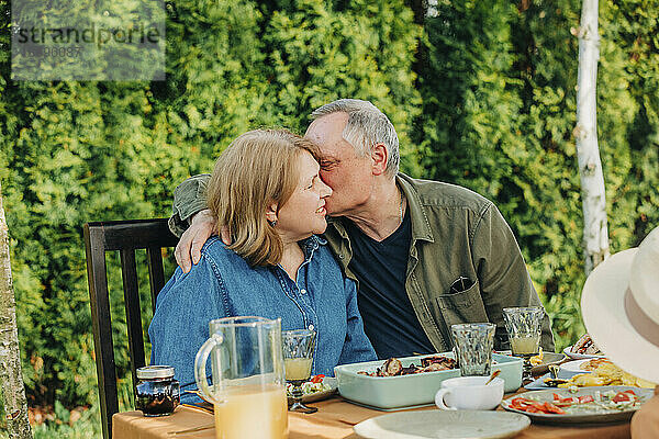 Älterer Mann küsst Frau  die am Esstisch im Hinterhof sitzt