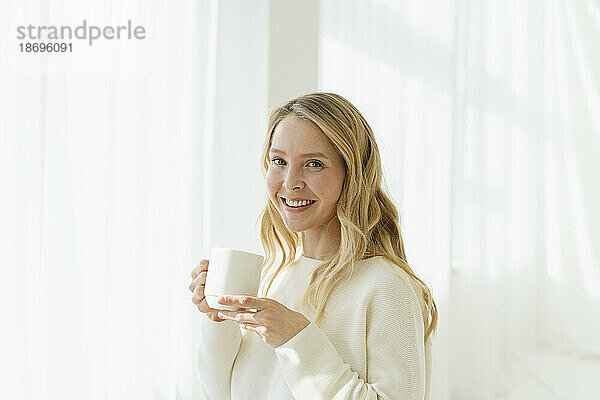 Lächelnde Frau hält Kaffeetasse vor durchscheinendem Vorhang