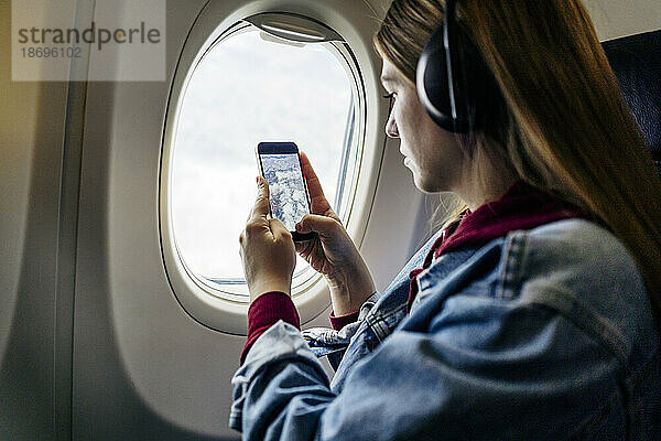 Frau mit Kopfhörern fotografiert mit Mobiltelefon durch Flugzeugfenster