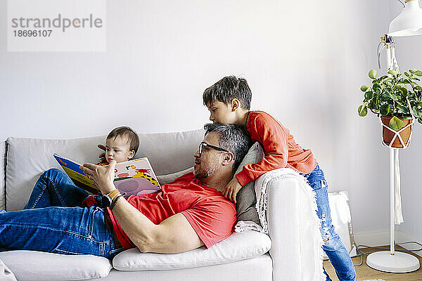 Vater liest Buch mit Söhnen auf Sofa im Wohnzimmer zu Hause