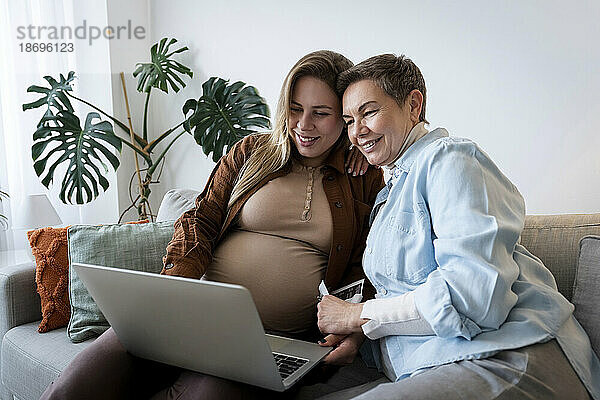 Lächelnde Frau beim Online-Shopping mit schwangerer Tochter über Laptop zu Hause