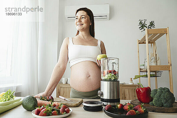 Lächelnde schwangere Frau bereitet zu Hause in der Küche Fruchtsmoothie zu