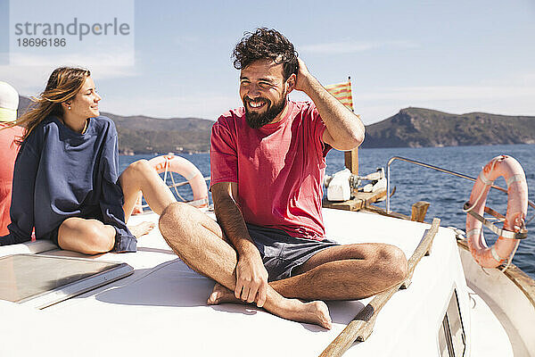 Lächelnder Mann sitzt im Schneidersitz neben Frau auf Yacht im Urlaub