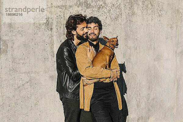 Gay man embracing boyfriend carrying dog in front of concrete wall