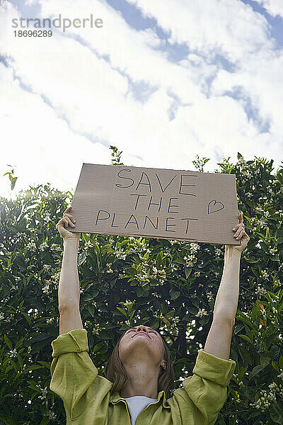 Frau hält einen ausgeschnittenen Karton mit dem Text „Save The Planet“ neben einem Baum im Garten