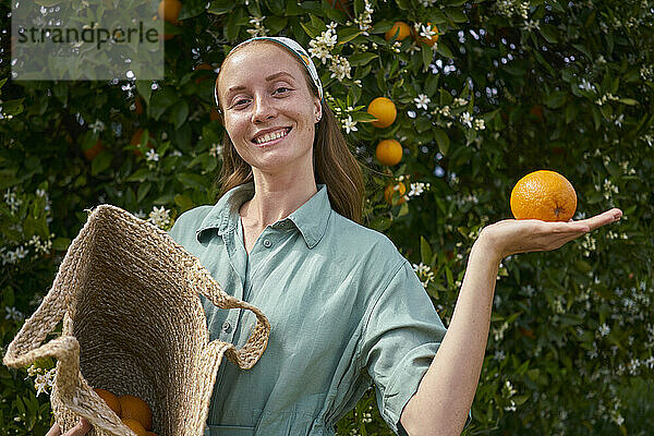 Lächelnde junge Frau mit Orange in der Hand neben Baum im Obstgarten