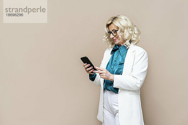Lächelnde Geschäftsfrau  die vor beigem Hintergrund mit ihrem Smartphone im Internet surft