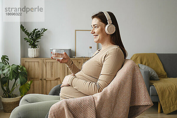 Lächelnde schwangere Frau  die zu Hause Musik hört und ein Glas Wasser in der Hand hält