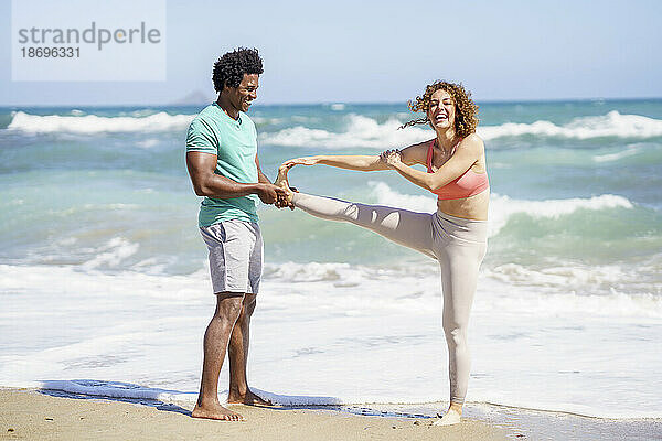 Mann hilft seiner Freundin beim Dehnen und Stehen auf einem Bein am Meeresufer