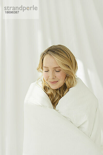 Lächelnde Frau mit geschlossenen Augen  in eine weiße Decke gehüllt  steht neben einem durchscheinenden Vorhang