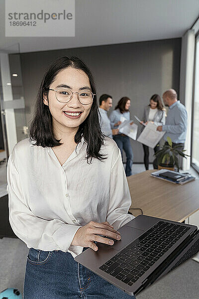 Porträt einer lächelnden jungen Geschäftsfrau  die einen Laptop mit Kollegen im Hintergrund im Büro benutzt
