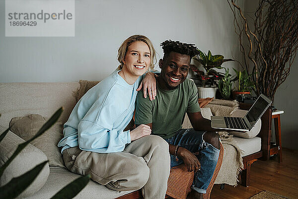 Lächelndes Paar sitzt mit Laptop im Wohnzimmer zu Hause