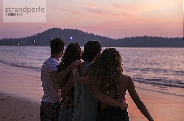 Freunde mit Armen stehen am Strand bei Sonnenuntergang