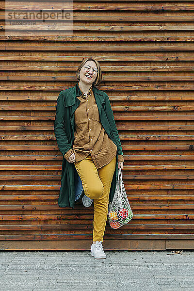 Lächelnde Frau im grünen langen Mantel lehnt an Fensterladen