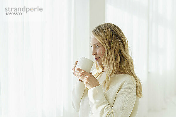 Lächelnde Frau trinkt Kaffee vor einem durchscheinenden Vorhang