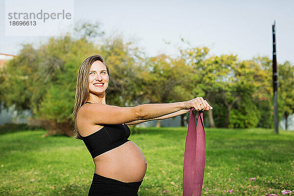 Lächelnde schwangere Frau trainiert mit Widerstandsband auf dem Rasen