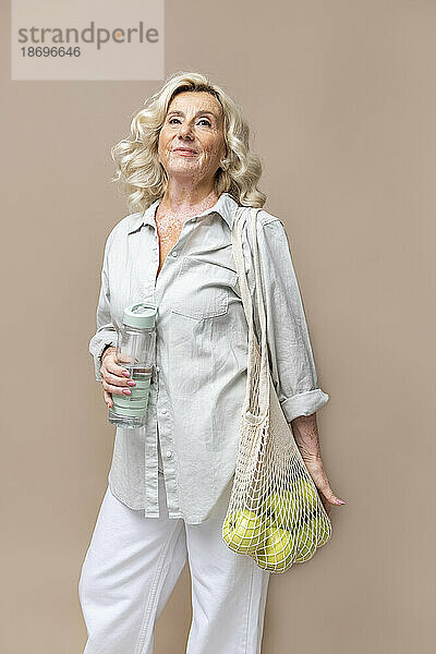 Ältere Geschäftsfrau mit Wasserflasche und Früchten im Netzbeutel vor beigem Hintergrund