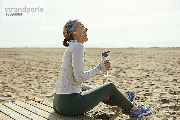 Frau mit Wasserflasche sitzt und lacht am Strand