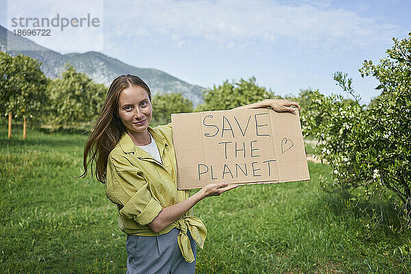 Lächelnde Frau zeigt den Text „Save The Planet“ auf einem im Garten ausgeschnittenen Karton