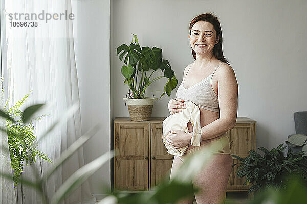 Smiling pregnant woman holding baby clothes at home