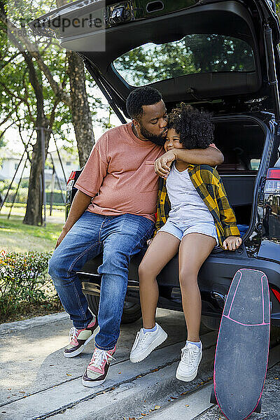 Father kissing daughter sitting on car trunk