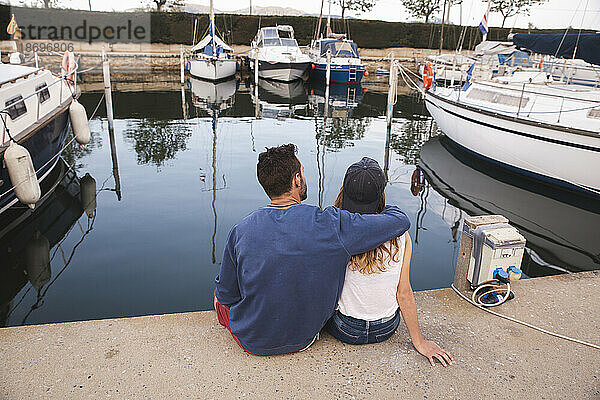 Mann mit Arm um Freundin sitzt auf Steg am Hafen