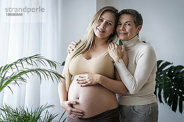Lächelnde ältere Frau umarmt schwangere Tochter  die zu Hause steht