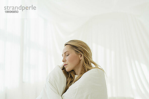 Frau mit geschlossenen Augen  eingehüllt in eine weiße Decke und durch einen durchscheinenden Vorhang