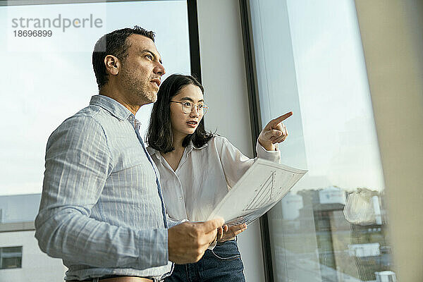 Geschäftsmann und Geschäftsfrau mit Architekturplan  der aus dem Fenster schaut