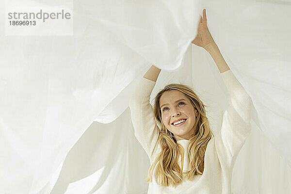Lächelnde Frau mit blonden Haaren  die mit einem weißen  durchscheinenden Vorhang spielt