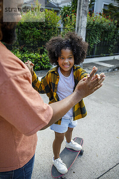 Mädchen steht auf Skateboard und hält die Hände ihres Vaters
