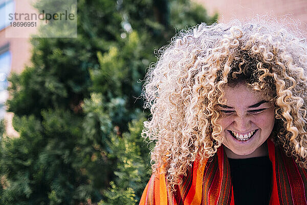 Young woman with curly hair laughing in front of tree