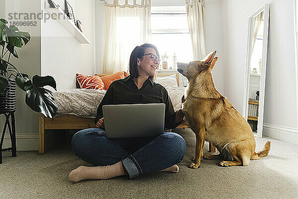 Freiberufler mit Laptop sitzt im Schneidersitz neben Hund zu Hause