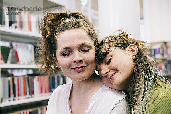 Lächelnde Frau stützt sich in der Bibliothek mit geschlossenen Augen auf eine Freundin