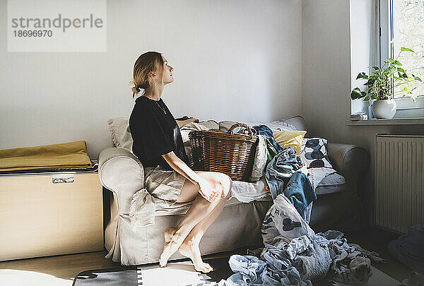 Nachdenkliche Frau sitzt zu Hause auf dem Sofa mit vollgestopfter Wäsche