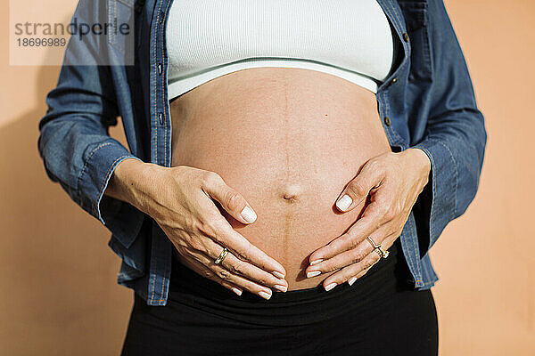 Schwangere Frau mit den Händen auf dem Bauch vor der Wand