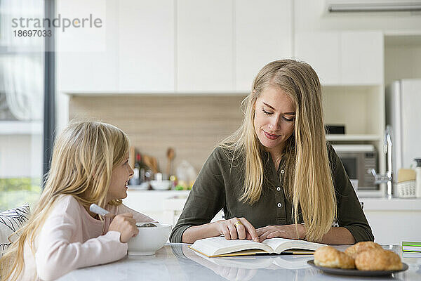 Mutter liest Buch und sitzt mit Tochter zu Hause in der Küche