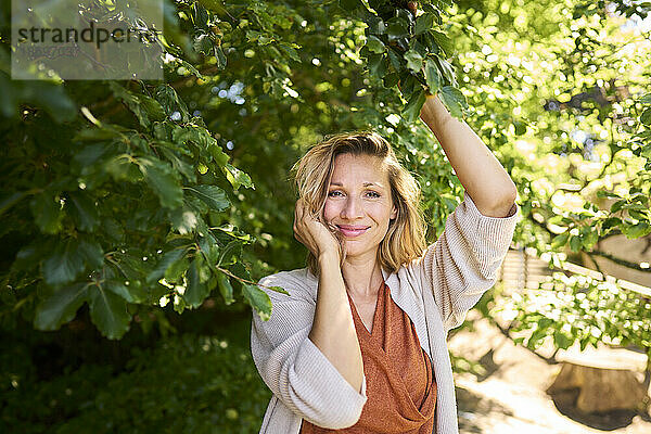 Lächelnde Frau mit der Hand im Haar  die unter einem Baum steht