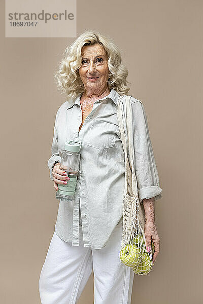 Senior businesswoman with mesh bag and water bottle against beige background