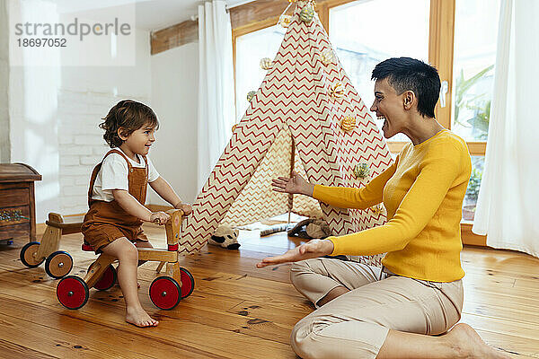 Fröhliche Frau spielt mit ihrem Sohn  der zu Hause auf dem Fahrrad sitzt