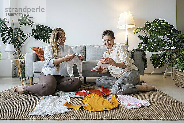 Glückliche Mutter und schwangere Tochter schauen sich die Babykleidung an  die zu Hause sitzt